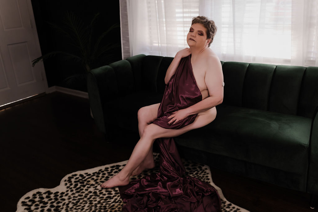 A woman with short hair sits on the edge of a couch draped in a maroon blanket in front of a window living health day spa