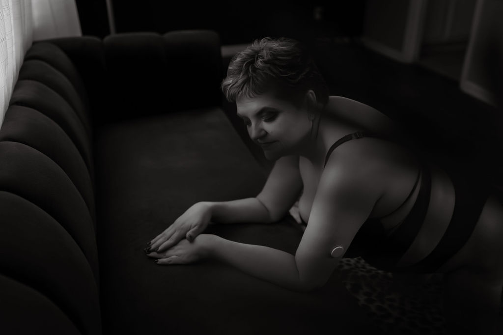 A woman in black lingerie kneels on the floor leaning onto a couch