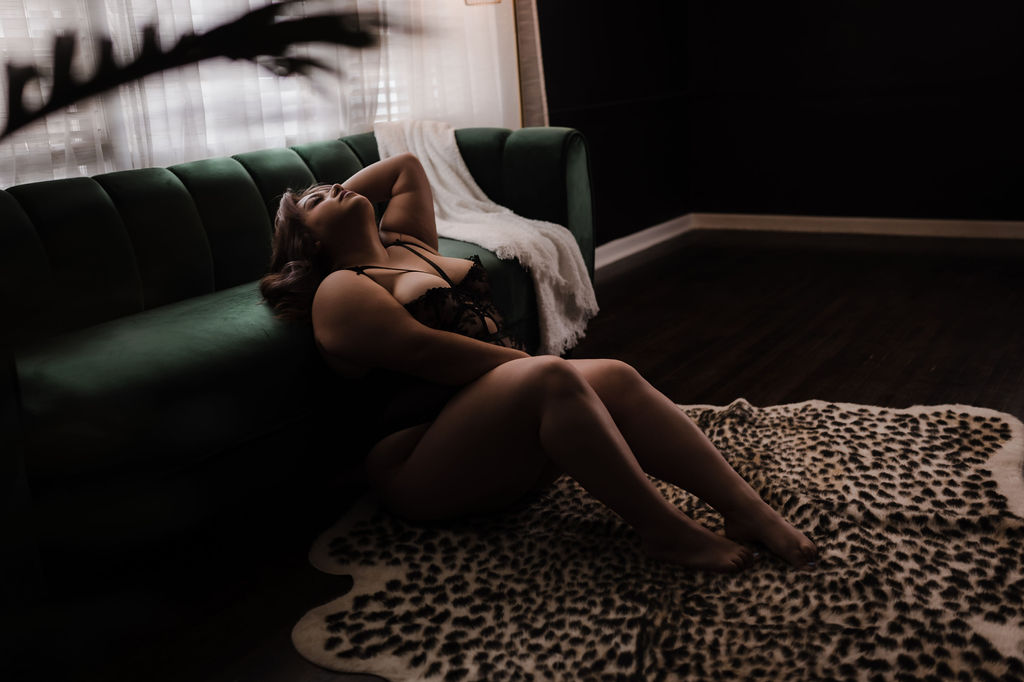 A woman in black lingerie sits on a leopard rug leaning against a couch