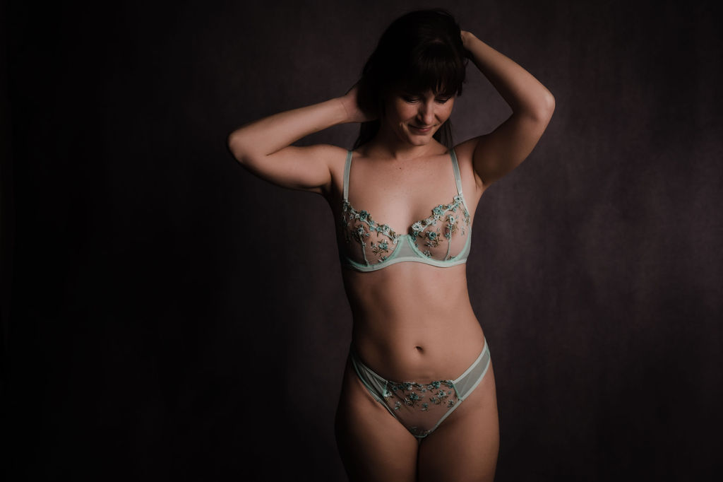 A woman in teal lingerie stands in a studio with her hands in her hair