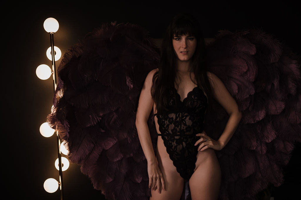 A woman in black lace lingerie and a feather backpack stands by a lamp in a studio after getting a brazillian wax memphis