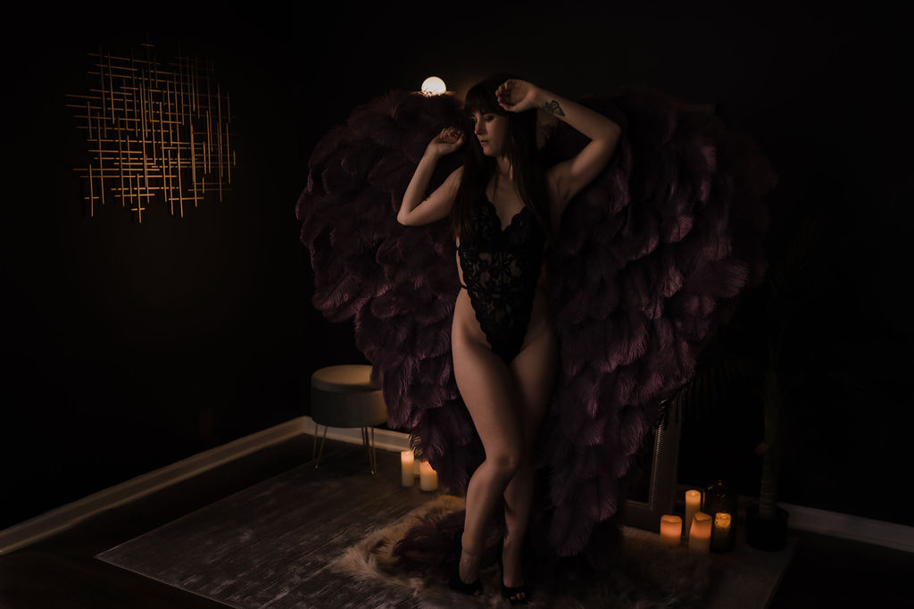 A woman in black lace lingerie and purple feather wings stands in a studio with candles after visiting seductions lingerie