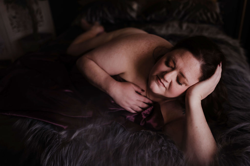 A woman lays across a dark bed covering herself with a purple silk sheet after microblading jonesboro ar